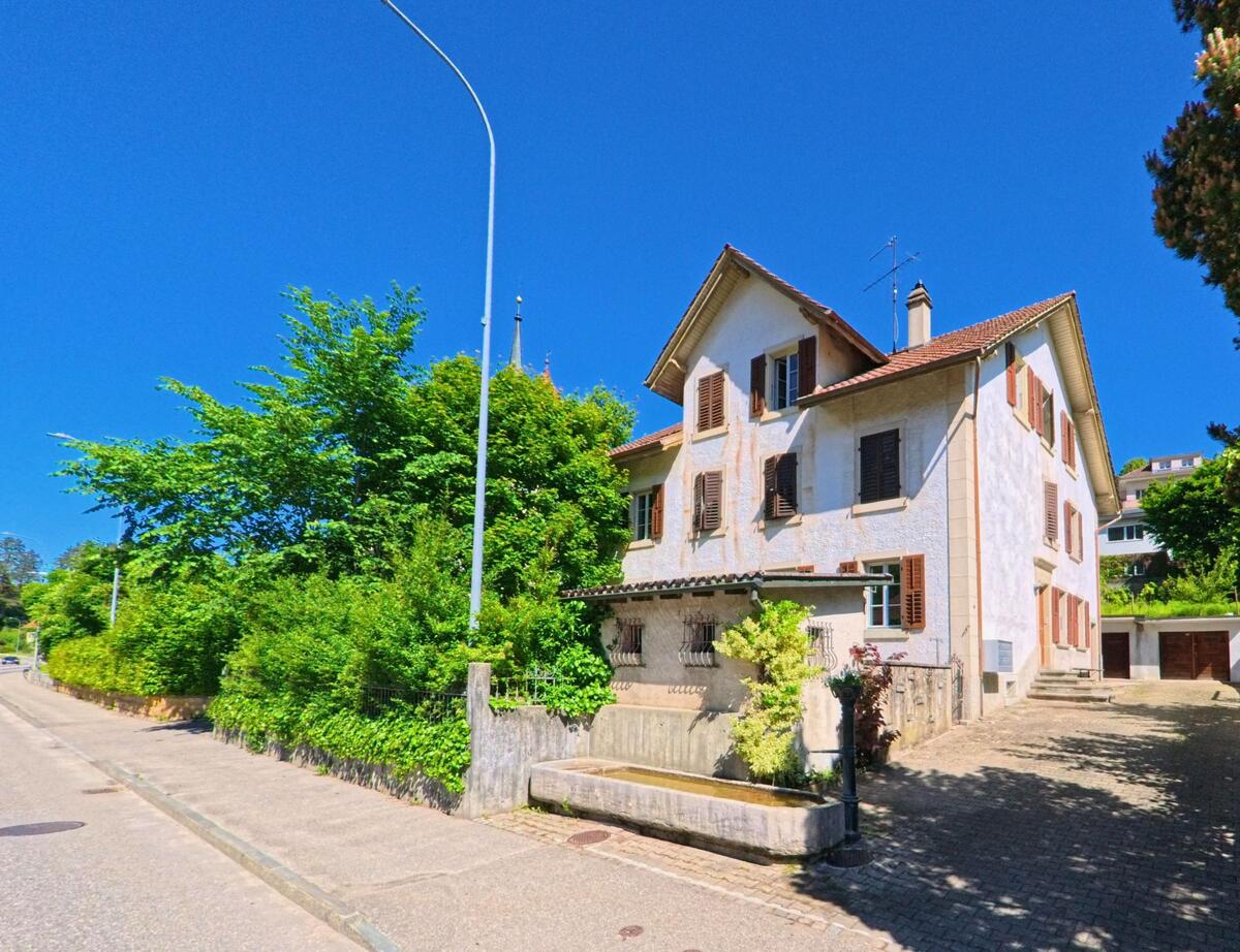 In Court (2738, Kanton Bern) steht ein helles Wohnhaus mit braunen Fensterläden direkt an einer Strasse mit traditionellem Brunnen im Vorgarten.