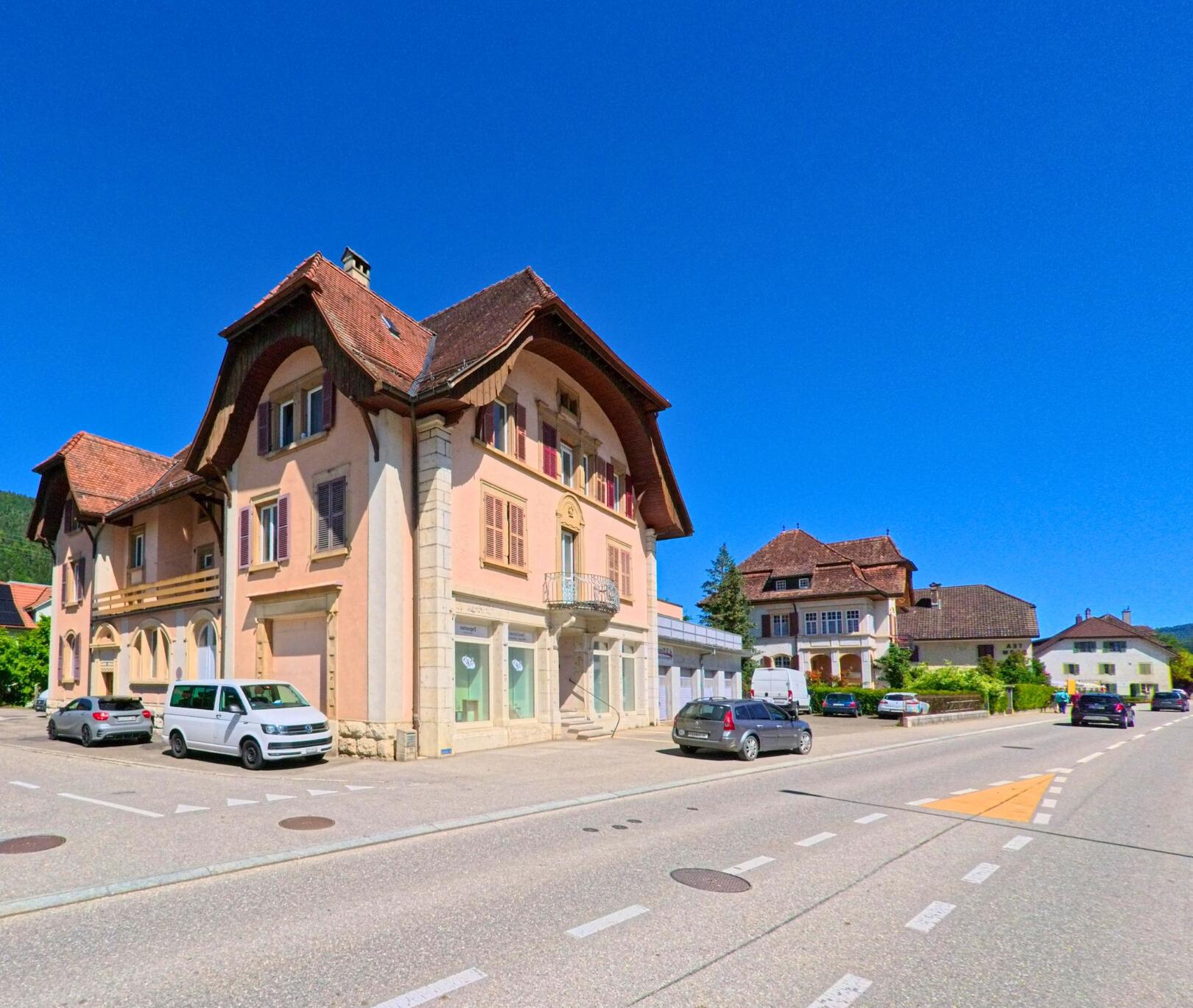 In Courtelary (2608, Kanton Bern) steht ein markantes Gebäude mit geschwungenem Dach und dekorativem Balkon direkt an der Hauptstrasse.