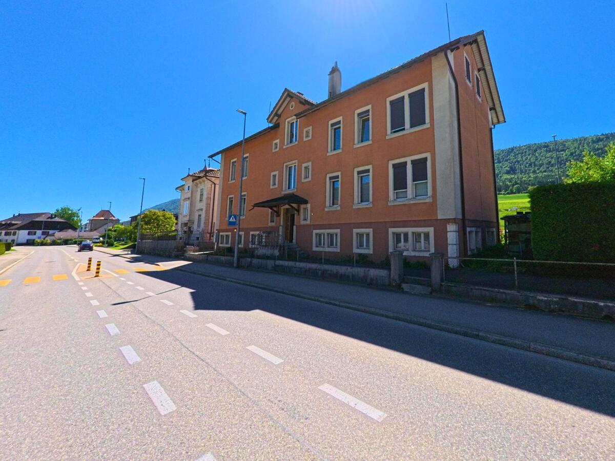 Entlang einer breiten Strasse in Courtelary (2608, Kanton Bern) reihen sich mehrstöckige Wohnhäuser mit Blick auf die angrenzende Hügellandschaft.