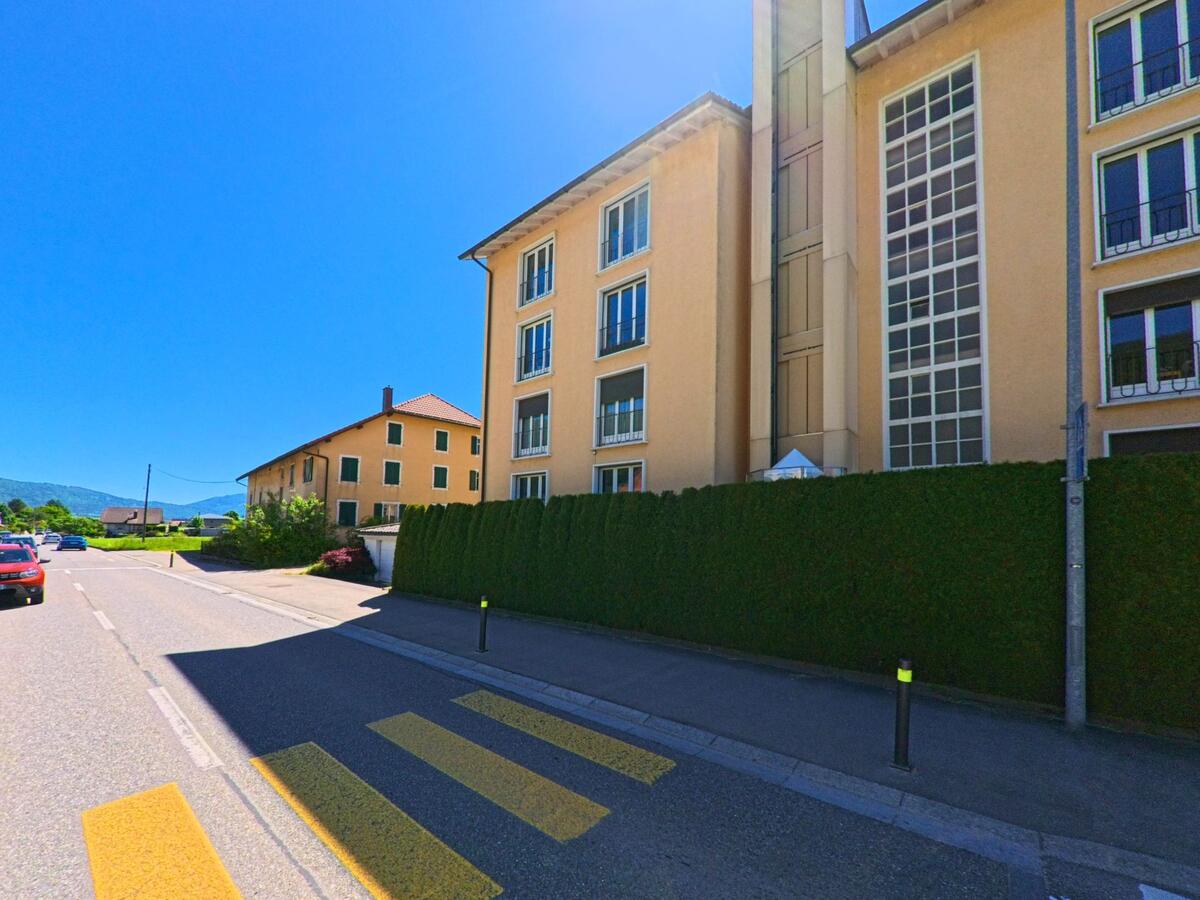 Entlang einer Hauptstrasse in Courtelary (2608, Kanton Bern) steht ein modernes Gebäude mit grosser Fensterfassade neben einem gepflegten Sichtschutz.