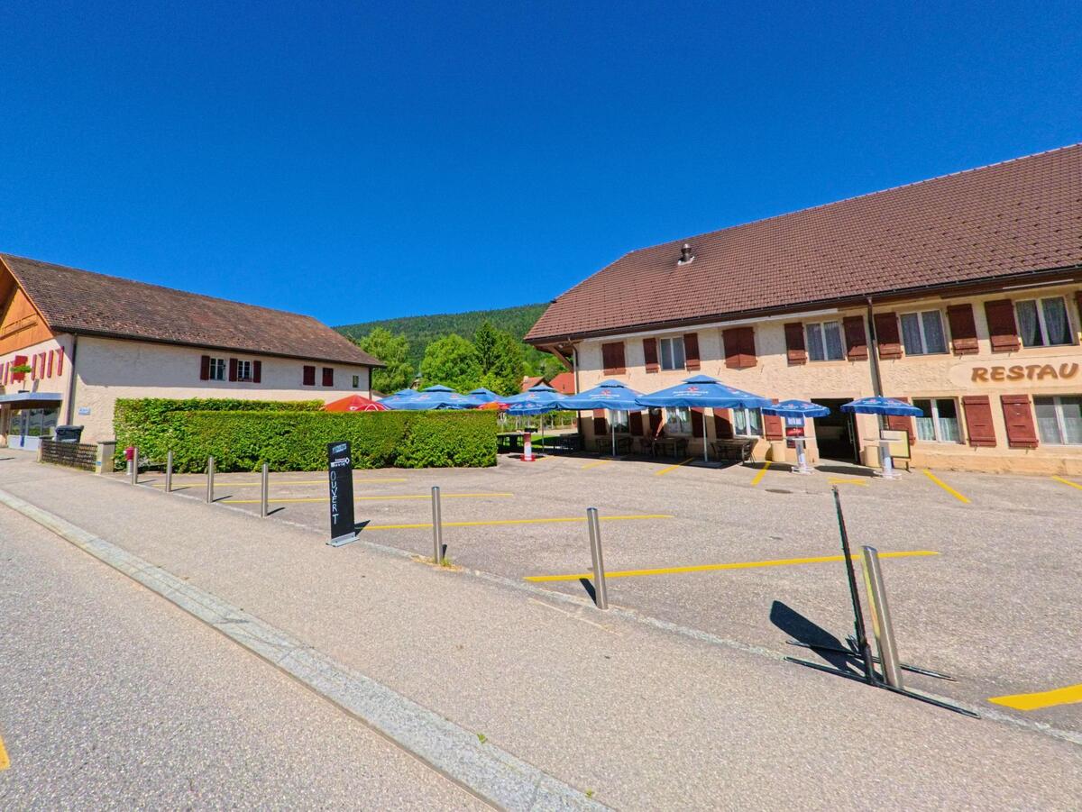In Courtelary (2608, Kanton Bern) lädt eine Terrasse mit Sonnenschirmen vor einem Restaurantgebäude mit Fensterläden zum Verweilen ein.