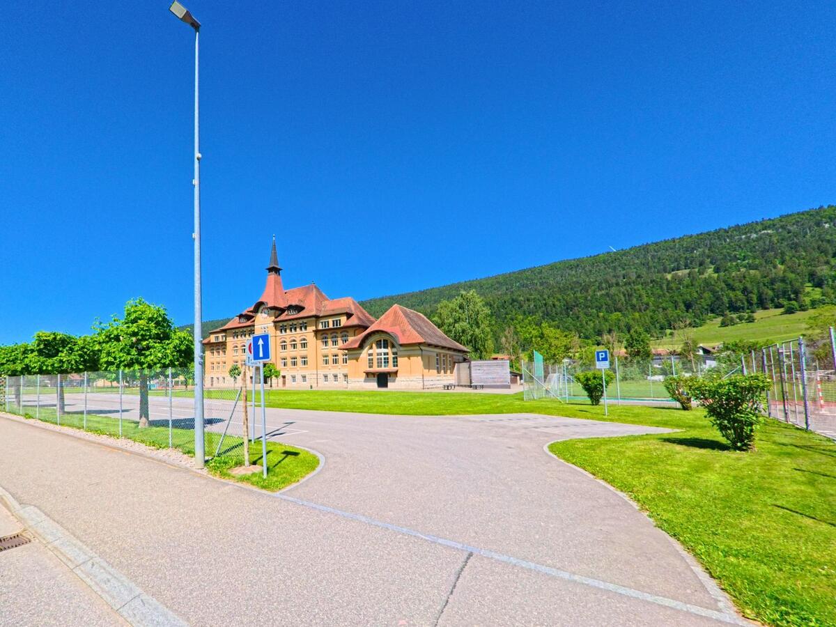 In Courtelary (2608, Kanton Bern) steht ein markantes Schulgebäude mit Türmchen und rotem Ziegeldach am Rand einer grossen Freifläche vor bewaldetem Hang.