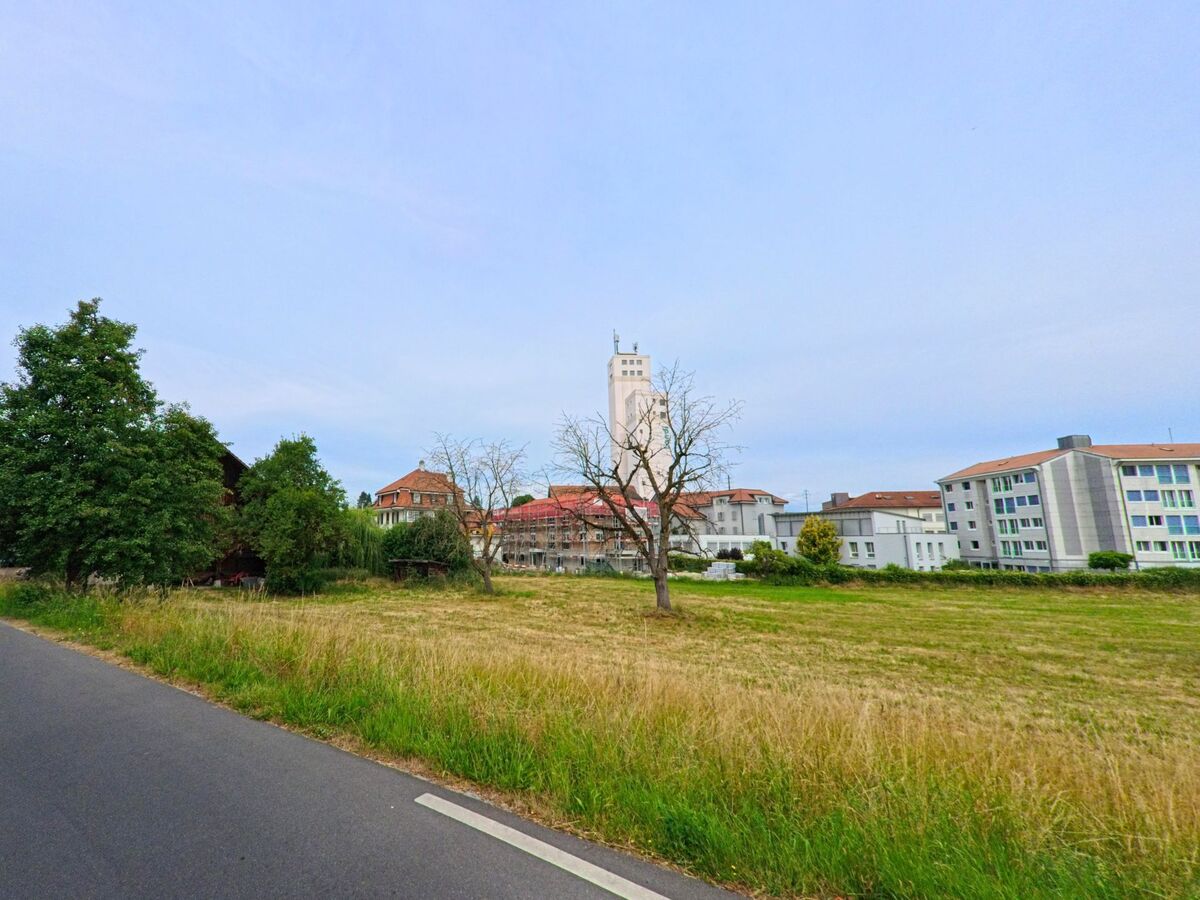 In Courtepin (1784, Kanton Freiburg) verläuft eine asphaltierte Strasse entlang eines Feldes mit Blick auf moderne Gebäude und einen markanten weissen Turm im Hintergrund.
