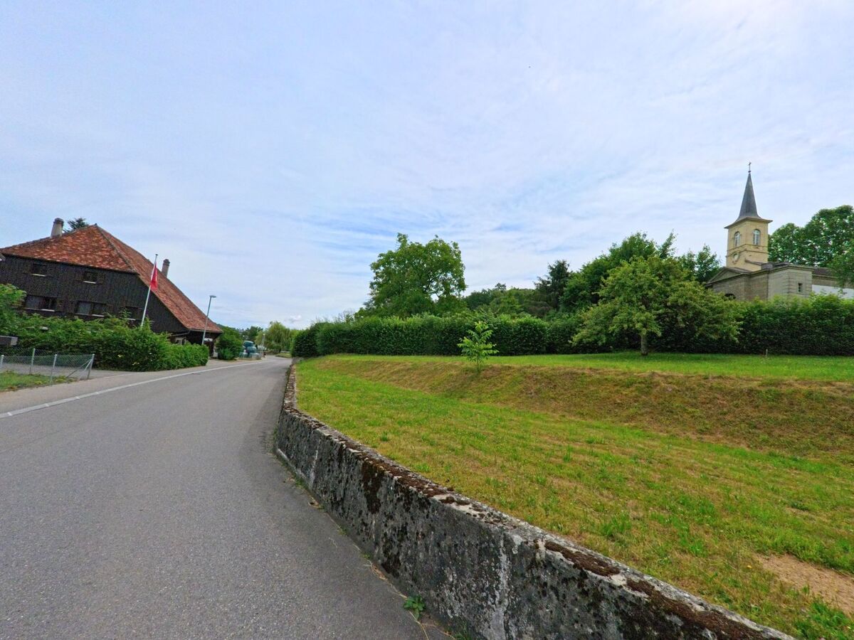 In Courtepin (1784, Kanton Freiburg) führt eine ruhige Strasse an einem alten Bauernhaus und einem erhöht gelegenen Kirchengebäude mit markantem Turm vorbei.