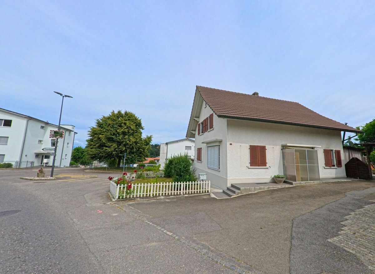In Courtepin (1791, Kanton Freiburg) steht ein Einfamilienhaus mit roten Fensterläden und kleinem Vorgarten nahe einer Kreuzung mit Mehrfamilienhäusern.