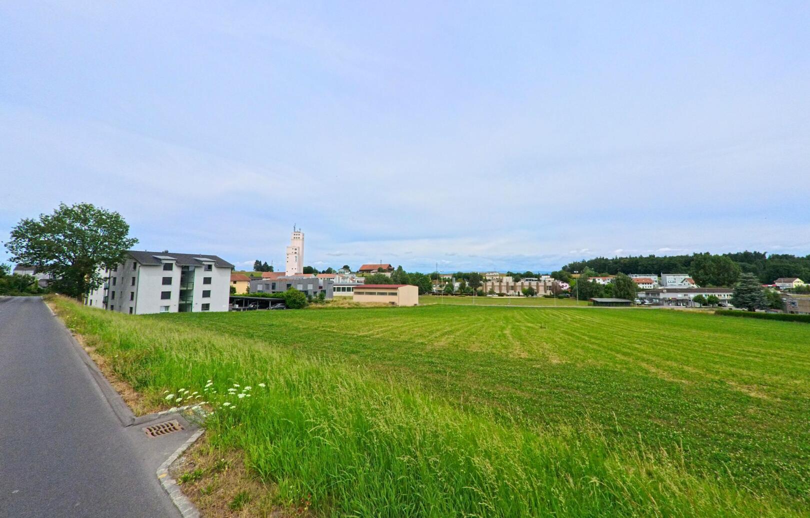 Eine asphaltierte Strasse in Courtepin (1791, Kanton Freiburg) verläuft entlang eines weiten Feldes mit Blick auf Wohngebäude, Gewerbebauten und einen hohen Turm in der Ferne.