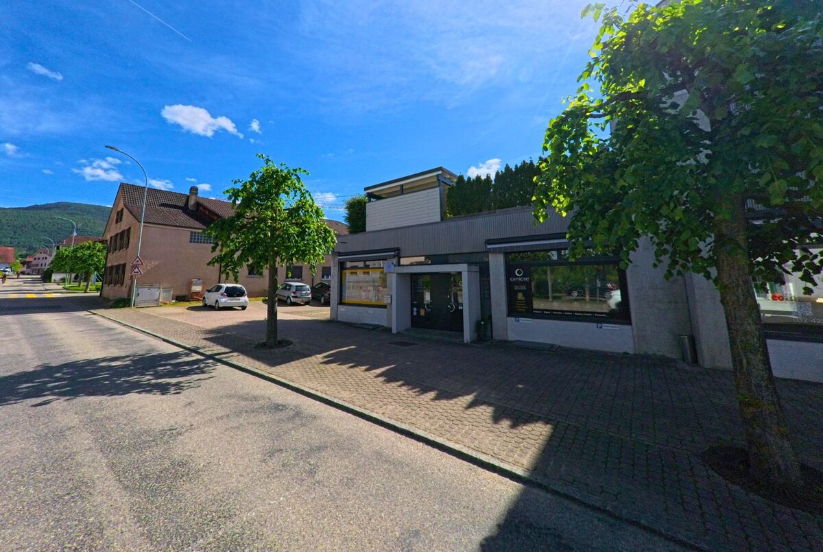 In Courtételle (2852, Kanton Jura) liegt eine moderne Gewerbeeinheit mit grossen Schaufenstern an einer ruhigen, von Bäumen gesäumten Strasse.