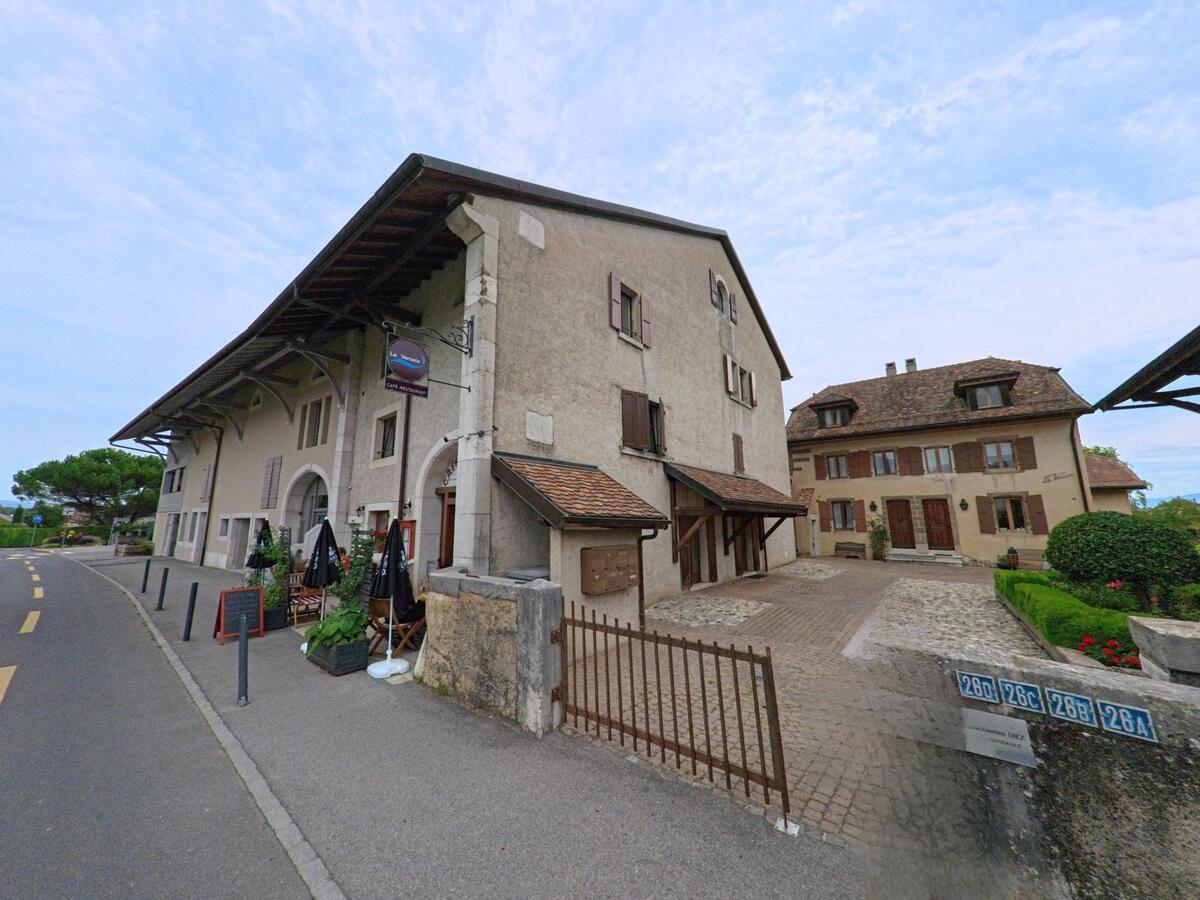 Ein Café in Crans (1299, Kanton Waadt) mit Tischen im Freien vor einem historischen Gebäude, während eine Person vorbeigeht und an einem Strassenschild liest.