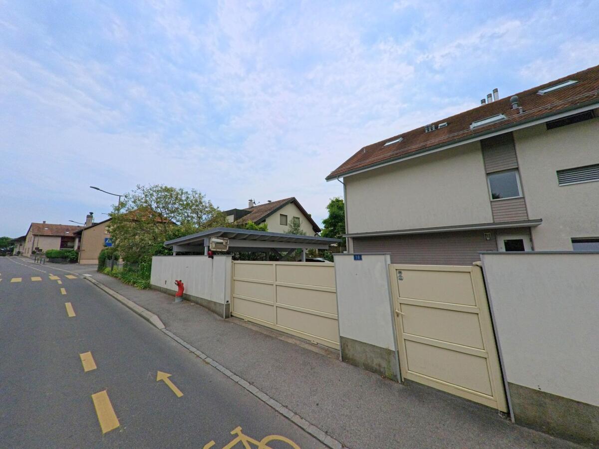 Ein modernes Haus mit einem weissen Tor und Carport an der Strasse in Crans (1299, Kanton Waadt), auf dem Gehweg sind Radwege markiert.
