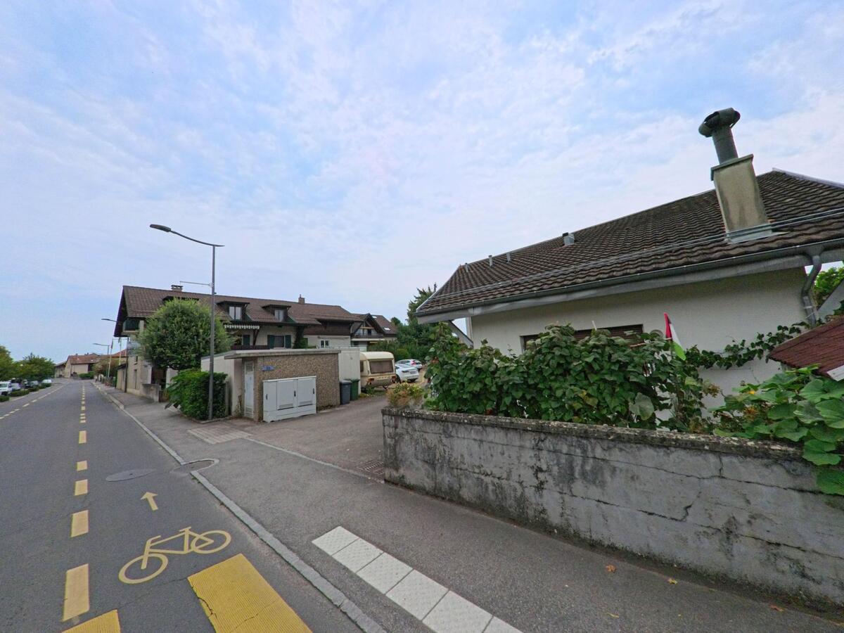 Ein Wohnhaus in Crans (1299, Kanton Waadt), mit einem gepflegten Gartenzaun und einem markierten Radweg entlang der Strasse.