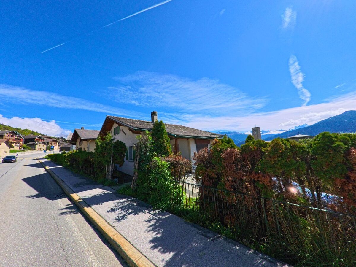 In Crans-Montana (3971, Kanton Wallis) stehen kleine Wohnhaeuser mit Gaerten an einer leicht ansteigenden Strasse, im Hintergrund sind Berge mit Schnee sichtbar.