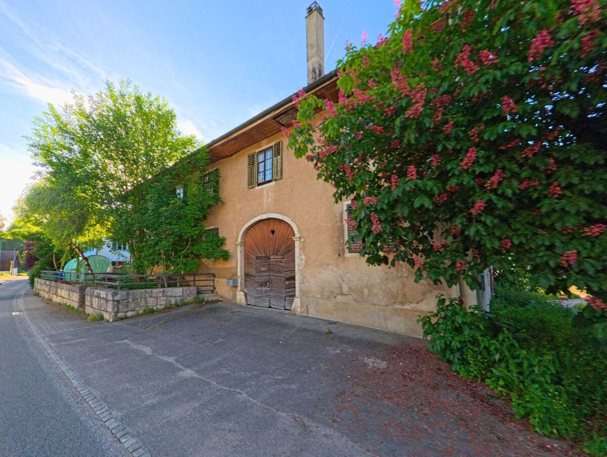 In Crémines (2746, Kanton Bern) steht ein altes Gebäude mit bogenförmigem Holztor und üppiger Blütenpracht an einem ruhig gelegenen Straßenabschnitt.