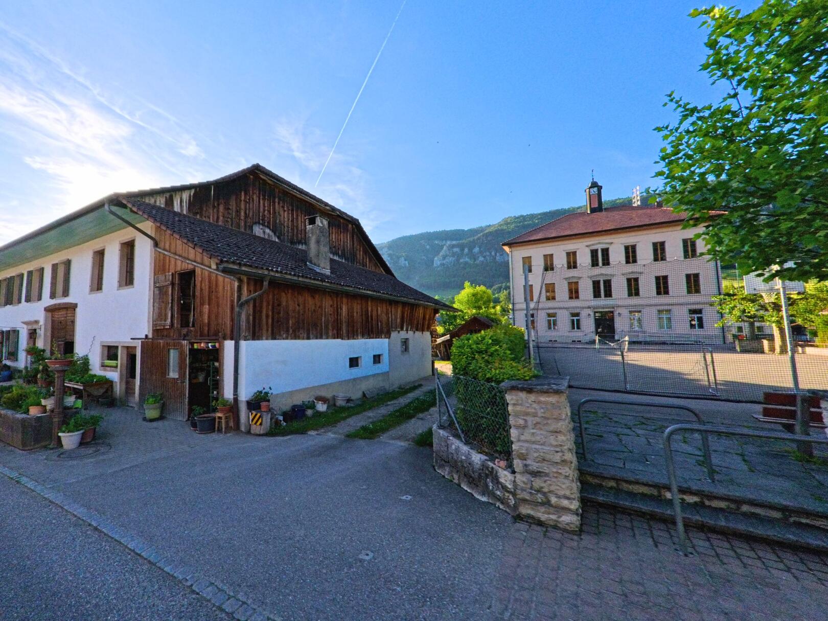 In Crémines (2746, Kanton Bern) steht ein traditionelles Bauernhaus gegenüber einem grossen, historischen Schulgebäude mit Pausenplatz.