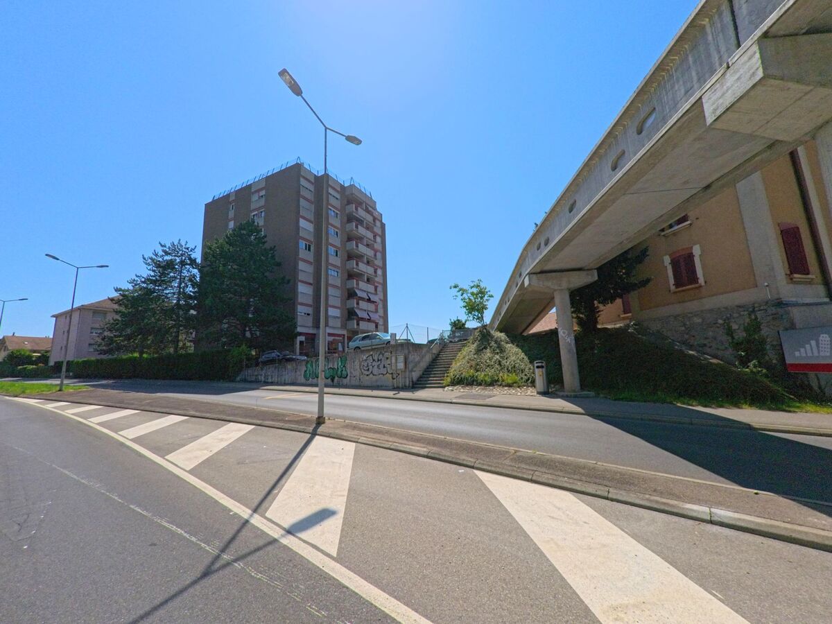 In Crissier (1023, Kanton Waadt) verläuft eine erhöhte Betonbrücke über einer Strasse mit Zebrastreifen und Strassenlaternen. Daneben steht ein hohes Wohnhaus, das den urbanen Charakter des Quartiers unterstreicht.