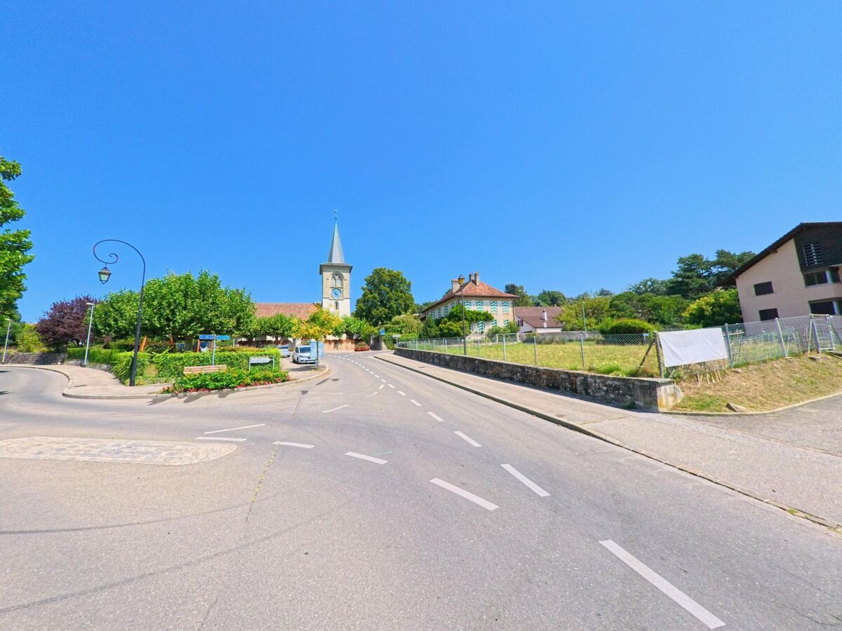 Eine Kirche mit spitzem Turm und Uhr erhebt sich im Ortszentrum von Crissier (1023, Kanton Waadt). Die Strasse führt leicht ansteigend auf das Gelände mit grünen Bäumen und historischen Gebäuden zu.