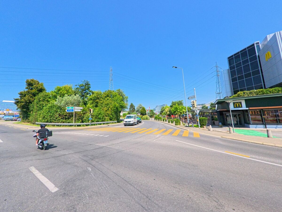 An einer Kreuzung in Crissier (1023, Kanton Waadt) liegt ein modernes Fast-Food-Restaurant mit markanter Glasfassade neben der Hauptstrasse. Ein Rollerfahrer und weitere Fahrzeuge passieren den Fussgängerstreifen bei strahlend blauem Himmel.