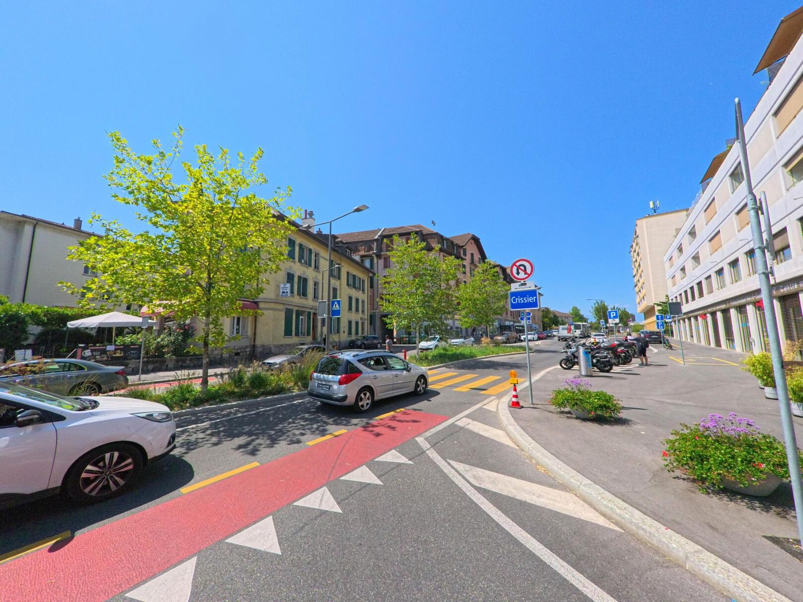 An der Einfahrt in Crissier (1023, Kanton Waadt) säumen Wohnhäuser, Geschäfte und parkende Fahrzeuge die Strasse. Das sonnige Wetter betont den Kontrast zwischen dem klaren Himmel und den grünen Bäumen entlang der Fahrbahn.