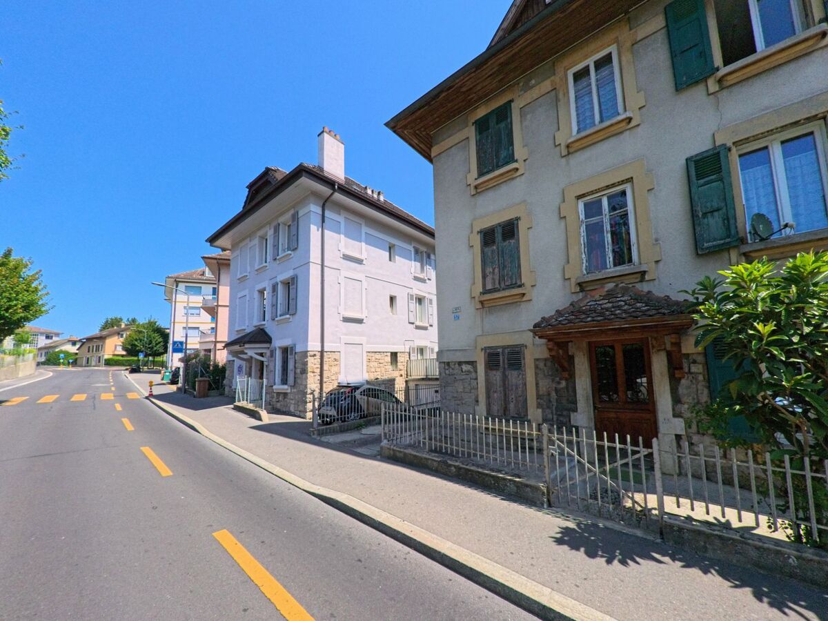 An einer leicht ansteigenden Strasse in Crissier (1023, Kanton Waadt) reihen sich ältere Wohnhäuser mit charakteristischen Fensterläden. Der asphaltierte Weg ist mit gelben Markierungen versehen und wird von Bäumen und modernen Gebäuden im Hintergrund begleitet.