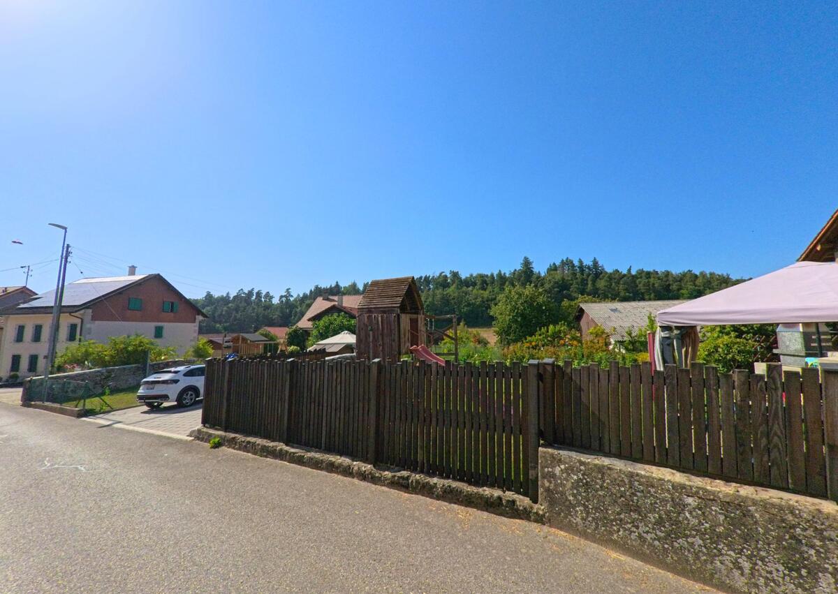 In Cronay (1406, Kanton Waadt) stehen Einfamilienhaeuser mit Gaerten entlang einer ruhigen Strasse, hinter einem Holzzaun ist ein Pavillon sichtbar.