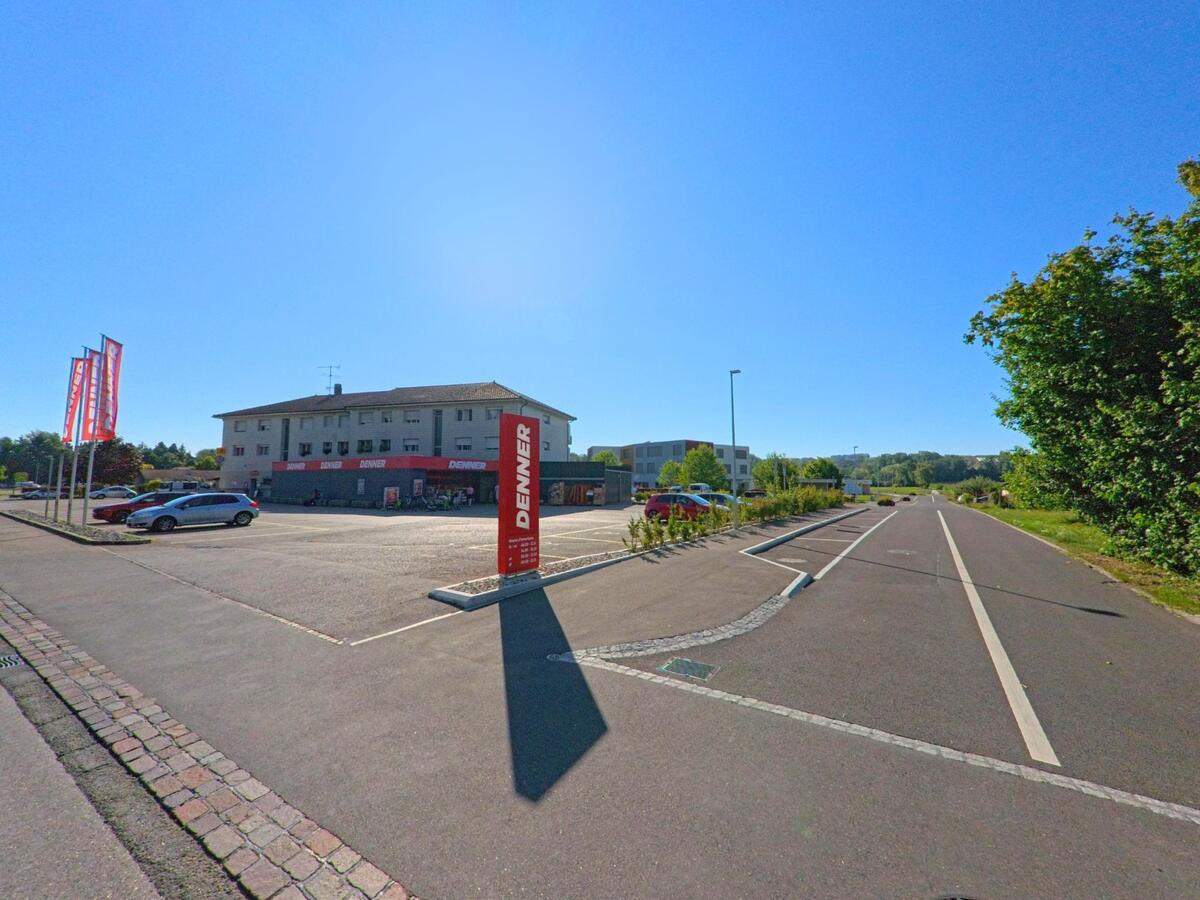 Unter strahlender Sonne liegt in Cudrefin (1588, Kanton Waadt) ein Denner-Supermarkt mit grossem Parkplatz an einer breiten Strasse mit Radweg.
