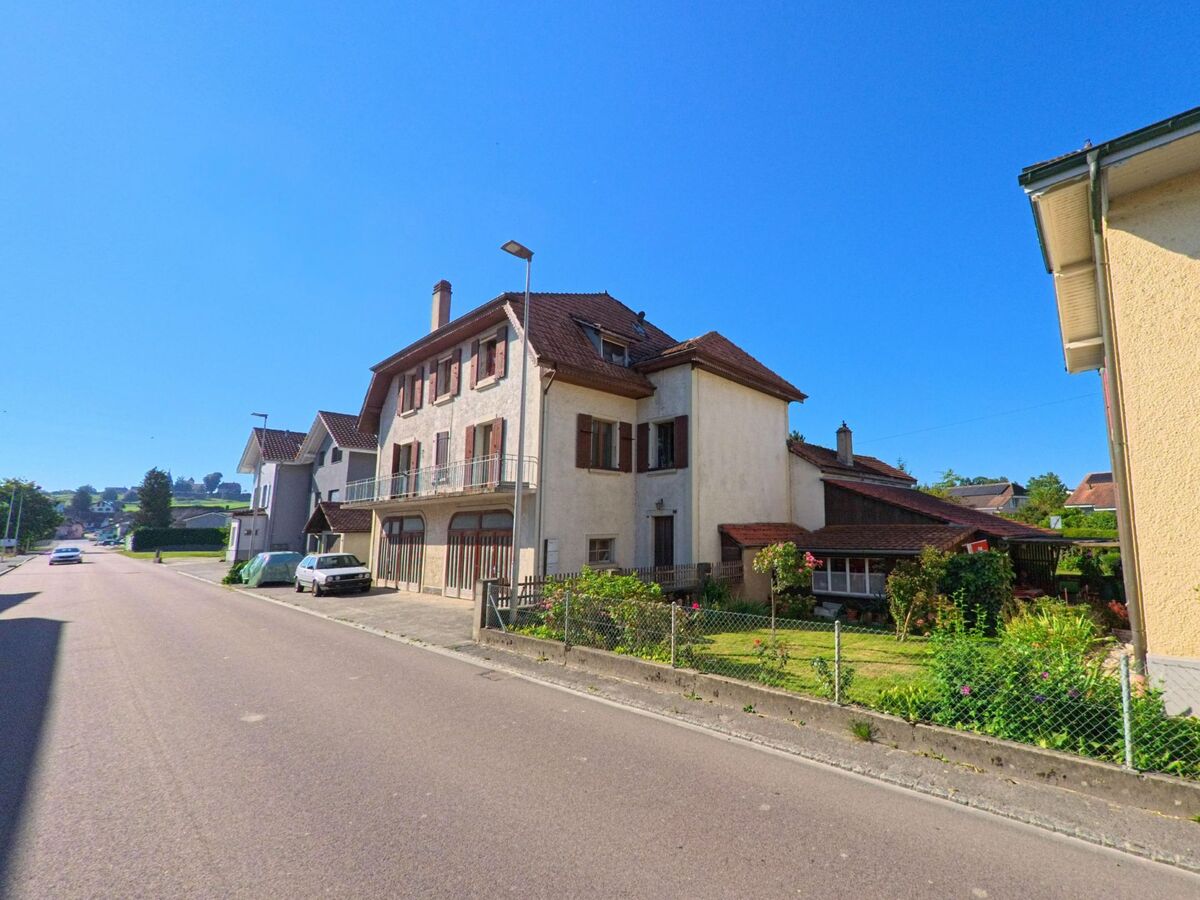 Entlang einer Strasse in Cudrefin (1588, Kanton Waadt) stehen Wohnhäuser mit Balkonen und gepflegten Vorgärten hinter niedrigen Zäunen.