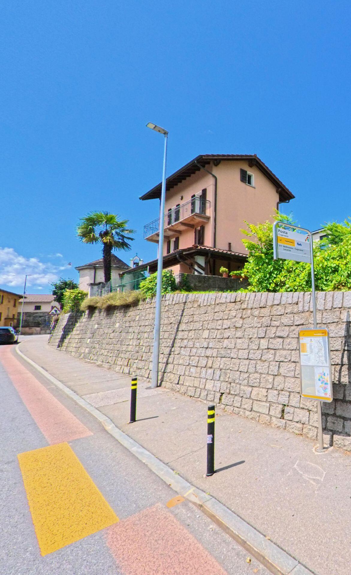 An einer Bushaltestelle in Cugnasco-Gerra (6516, Kanton Tessin) steht ein mehrstöckiges Haus mit Balkonen oberhalb einer Steinmauer unter klarem Himmel.