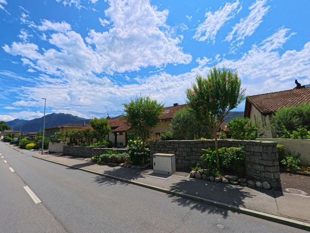Entlang einer gepflegten Strasse in Cugnasco-Gerra (6516, Kanton Tessin) stehen Einfamilienhäuser mit Gärten und Blumen vor der Bergkulisse.