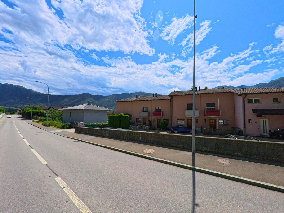 Reihenhäuser mit roten Balkonen säumen in Cugnasco-Gerra (6516, Kanton Tessin) eine breite Strasse mit Blick auf die umliegenden Berge.