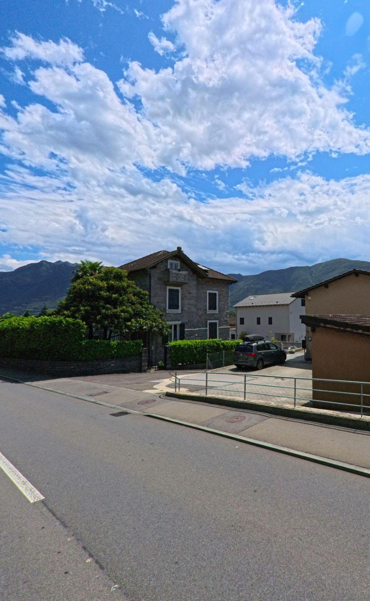 Ein Steinhaus mit Garten und Einfahrt steht in Cugnasco-Gerra (6516, Kanton Tessin) vor einer Kulisse aus Bergen und blauem Himmel.