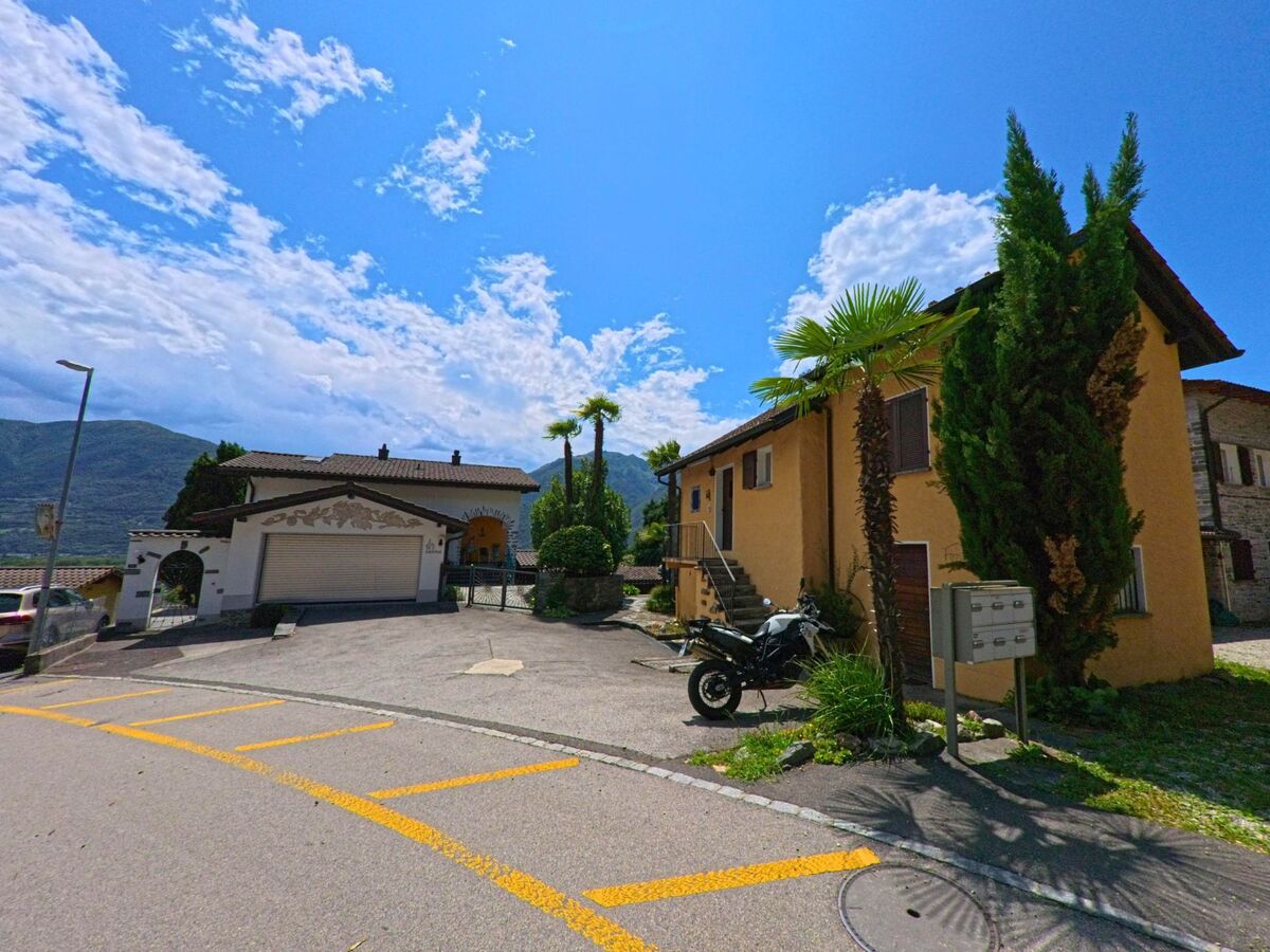Zwei Wohnhaeuser mit Palmen und Einfahrten liegen in Cugnasco-Gerra (6516, Kanton Tessin) an einer leicht ansteigenden Strasse mit Blick auf die Berge.