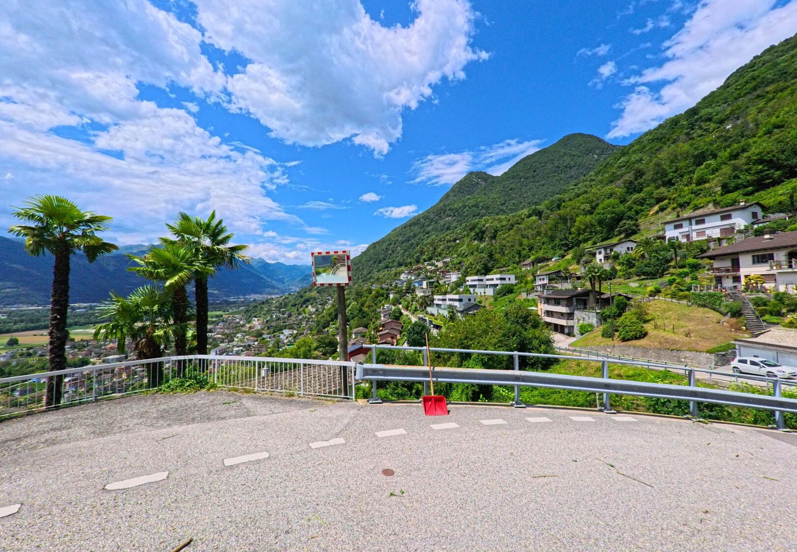 Von einer Aussichtskurve in Cugnasco-Gerra (6597, Kanton Tessin) bietet sich ein weiter Blick über das Tal mit Palmen, Hängen und den umliegenden Häusern.