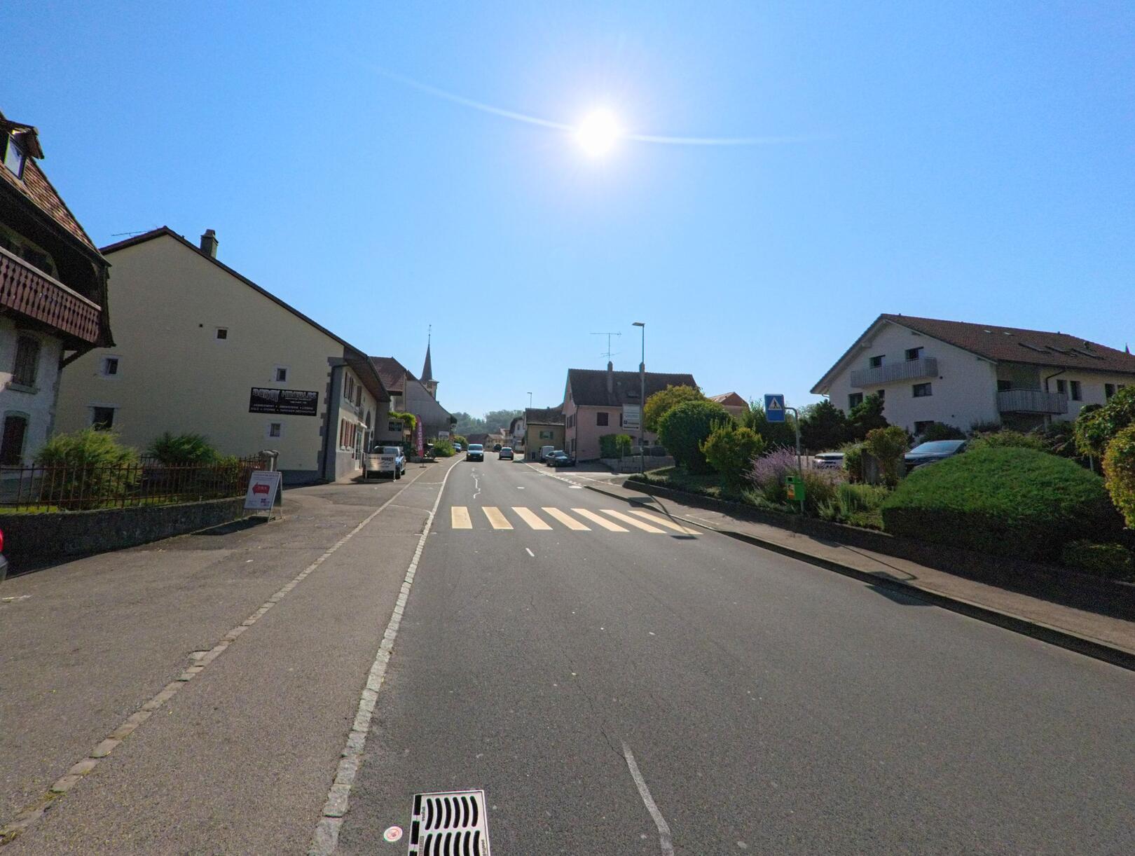 Eine Hauptstrasse in Cugy (1482, Kanton Freiburg) fuehrt durch den Ortskern, im Hintergrund ist der Kirchturm sichtbar.