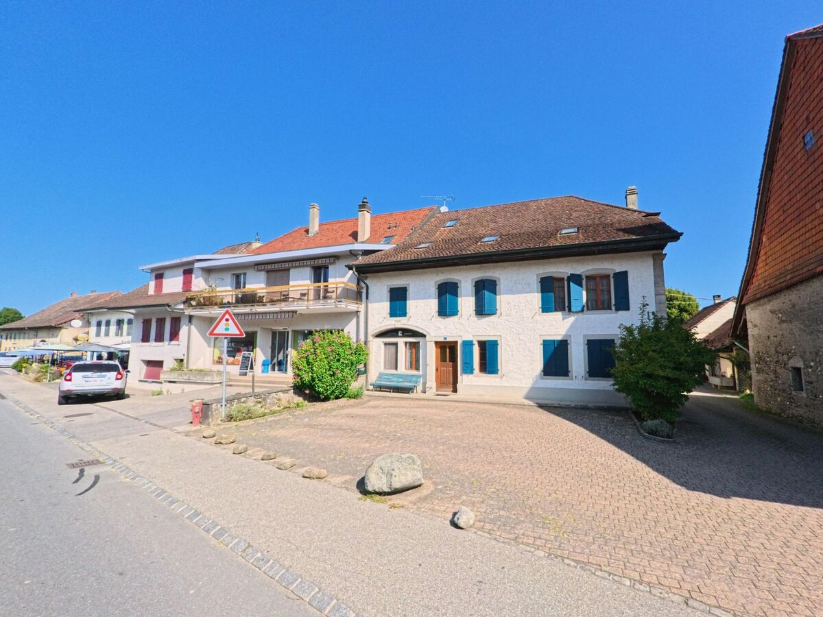 Mehrere traditionelle Wohnhaeuser mit farbigen Fensterlaeden und Balkonen stehen entlang einer Strasse in Cugy (1482, Kanton Freiburg).