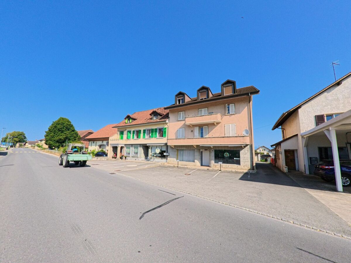 Ein Traktor faehrt die Strasse entlang, gesaeumt von Wohn- und Geschaeftshaeusern in Cugy (1482, Kanton Freiburg).