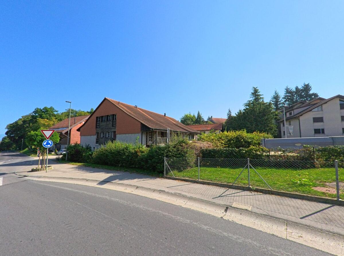 Ein traditionelles Bauernhaus mit rotem Dach steht an einer Strasse in Cugy (1483, Kanton Freiburg) neben modernen Wohngebaeuden.