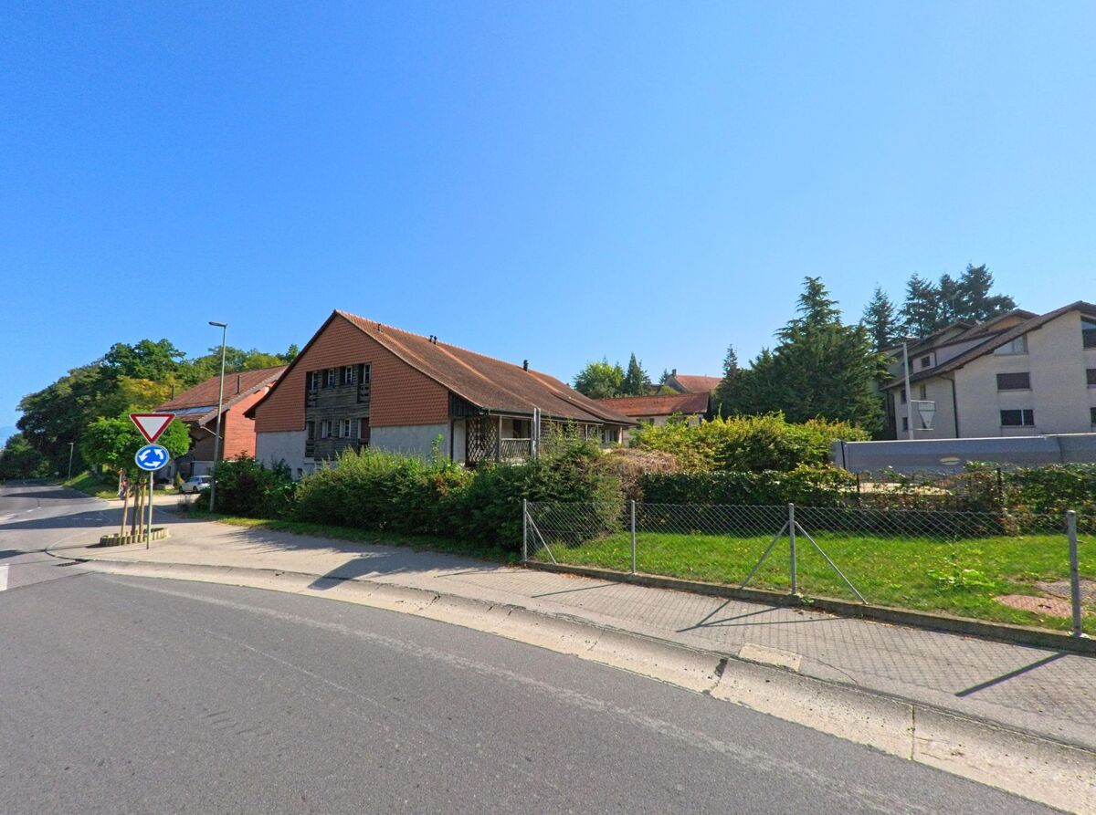 Ein traditionelles Bauernhaus mit rotem Dach steht an einer Strasse in Cugy (1483, Kanton Freiburg) neben modernen Wohngebaeuden.