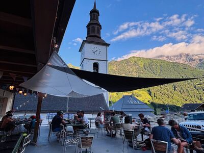 Auf der Terrasse des Culture(s) Café Cantine in Val-d’Illiez (1873, Kanton Wallis) geniessen Gäste bei sommerlichem Wetter den Blick auf die Kirche und die umliegenden Alpen.