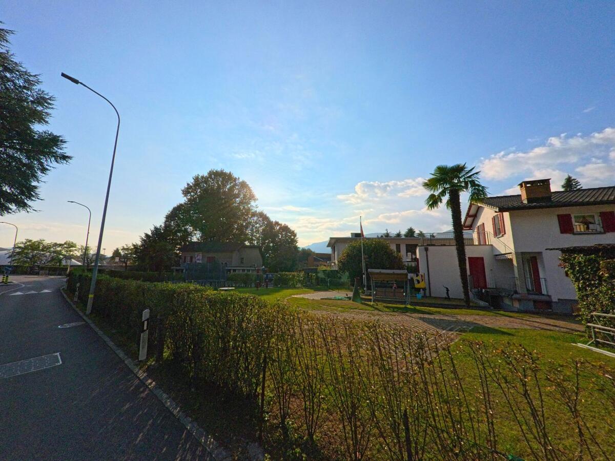 Neben einer ruhigen Strasse liegt eine grüne Freifläche mit Wohnhäusern, Bäumen und Hecken in Cureglia (6944, Kanton Tessin) im weichen Tageslicht.