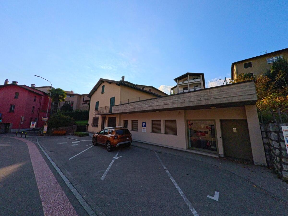 Vor einem niedrigen Gebäude mit Vordach und markierten Parkfeldern liegt ein kleiner Platz mit Wohnhäusern in Cureglia (6944, Kanton Tessin) unter klarem Himmel.