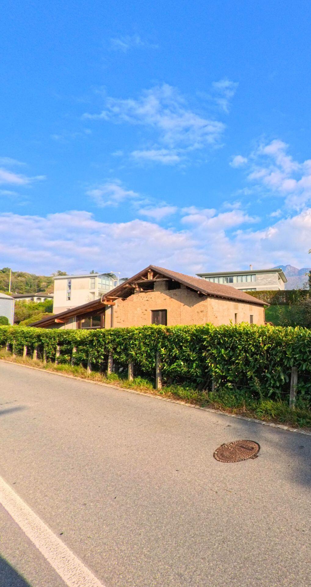 Hinter einer dichten Hecke steht ein niedriges, traditionelles Gebäude an einer Nebenstrasse in Cureglia (6944, Kanton Tessin) unter weitem Himmel.