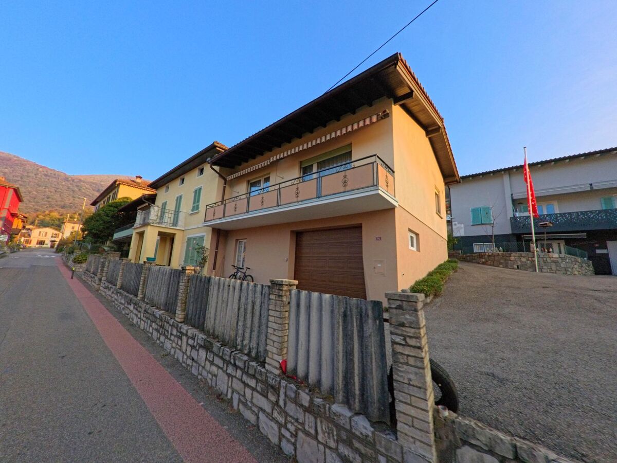 Entlang einer ruhigen Quartierstrasse stehen in Curio (6981, Kanton Tessin) Wohnhäuser mit Balkonen, Steinmauern und Blick auf die umliegenden Hügel.