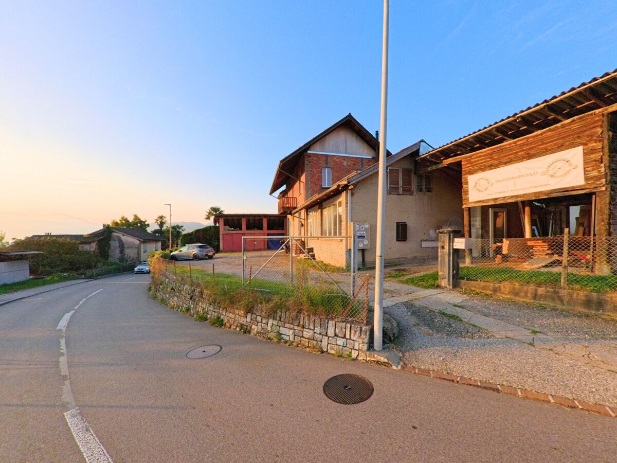 An einer leicht gebogenen Strasse in Curio (6984, Kanton Tessin) stehen ländliche Gebäude mit Hofzufahrt, Steinmauer und Werkbereich im warmen Abendlicht.