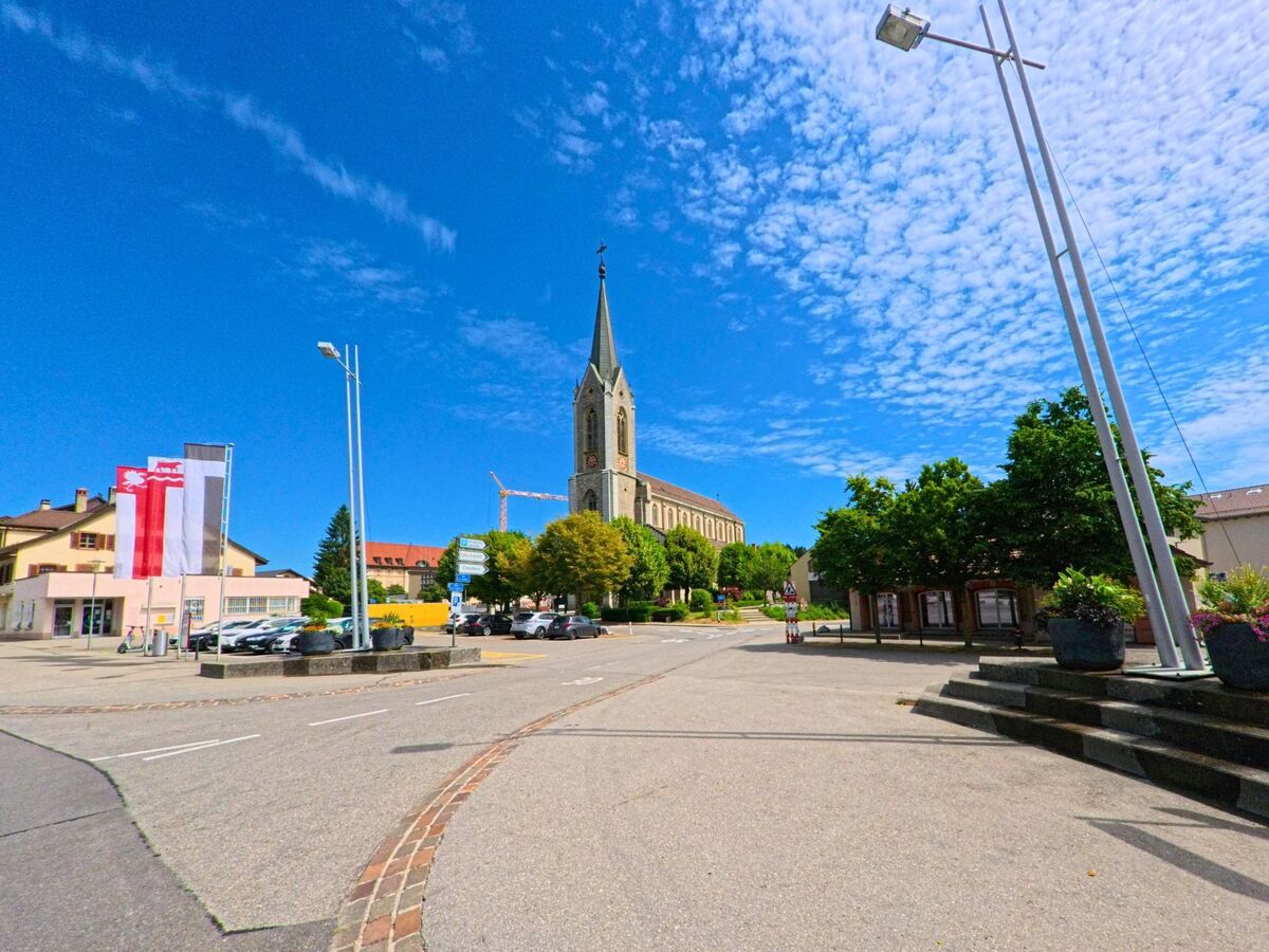 Der Blick auf die Kirche von Bulle (1635, Kanton Freiburg) eröffnet sich von einem weitläufigen Platz mit Fahnen, Wegweisern und parkenden Autos unter einem sommerlichen Himmel.