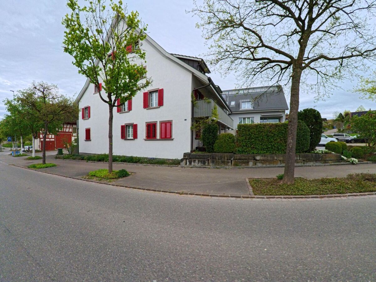 Ein Wohnhaus mit weisser Fassade und roten Fensterläden steht in Dägerlen (8442, Kanton Zürich) an einer baumbestandenen Strasse, flankiert von weiteren Wohnhäusern mit gepflegten Gärten.