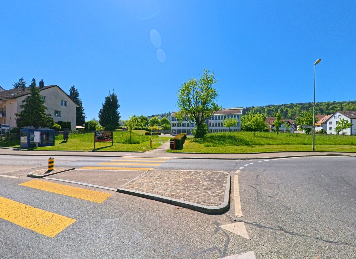 In Dällikon im Kanton Zürich mit der Postleitzahl 8108 führt ein Fussgängerstreifen über eine Strasse zu einer grosszügigen Grünfläche mit Weg und modernem Verwaltungsgebäude.