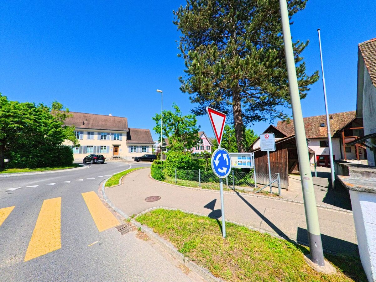 Ein kleiner Kreisel mit Verkehrszeichen liegt im Zentrum von Dällikon im Kanton Zürich, PLZ 8108, umgeben von historischen Gebäuden und Gehwegen.