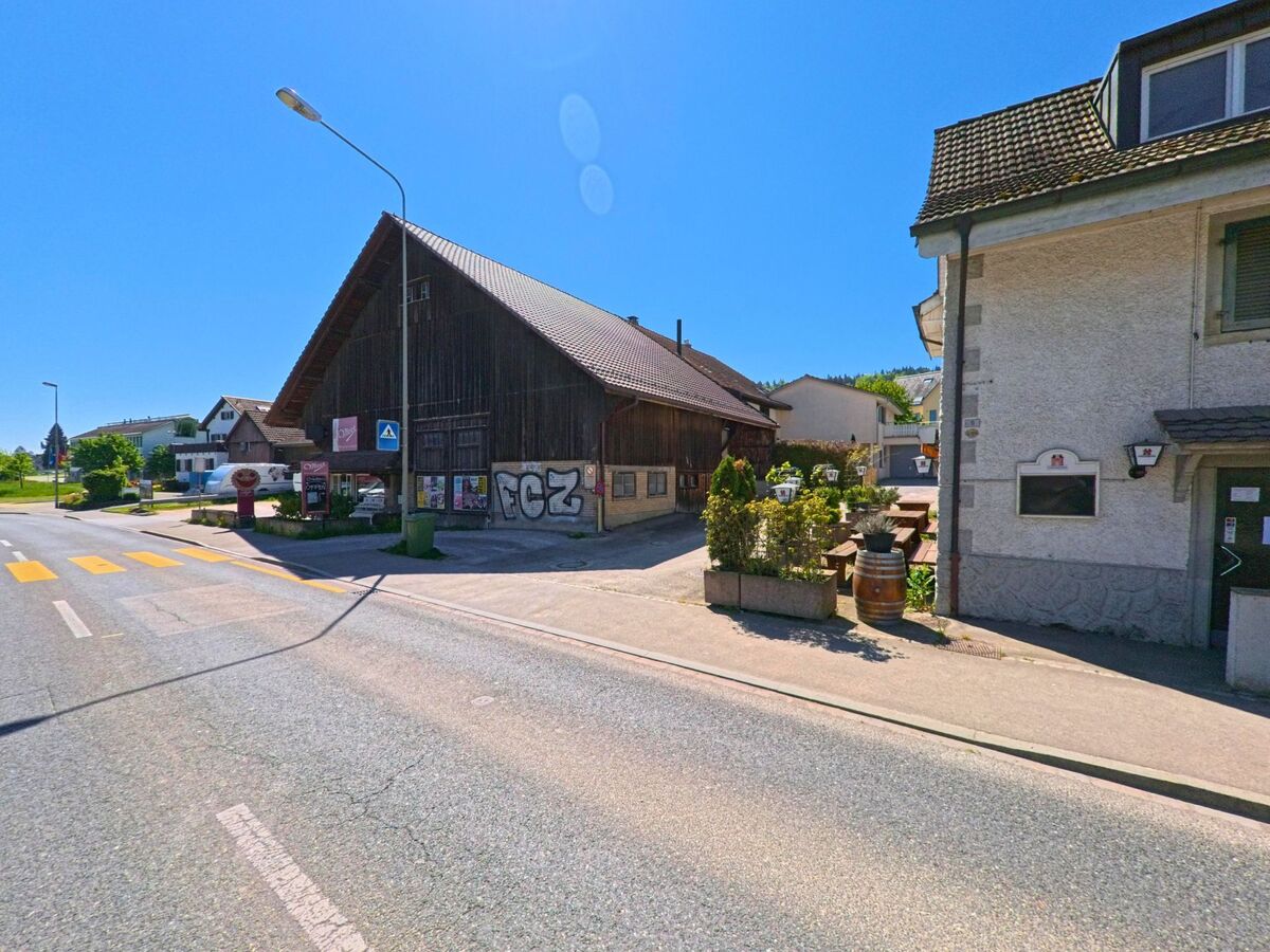 An einer Kreuzung in Dällikon im PLZ-Gebiet 8108 im Kanton Zürich steht ein grosses Holzgebäude mit Werbeplakaten und ein Restaurant mit Aussenplätzen.