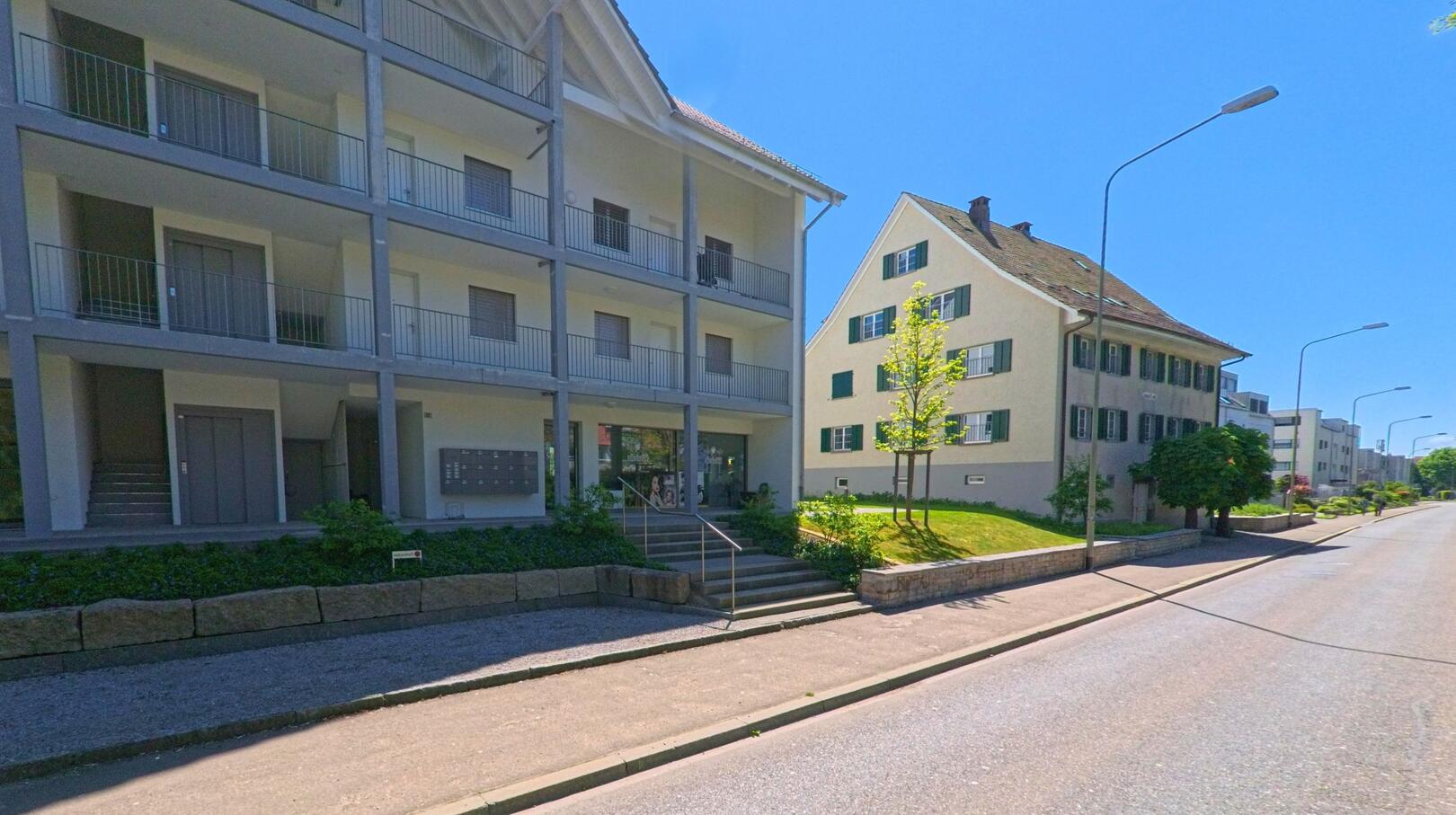 In Dällikon (8108, Kanton Zürich) stehen entlang einer ruhigen Strasse ein modernes Mehrfamilienhaus mit offenen Laubengängen und ein traditionelles Wohnhaus mit grünen Fensterläden.