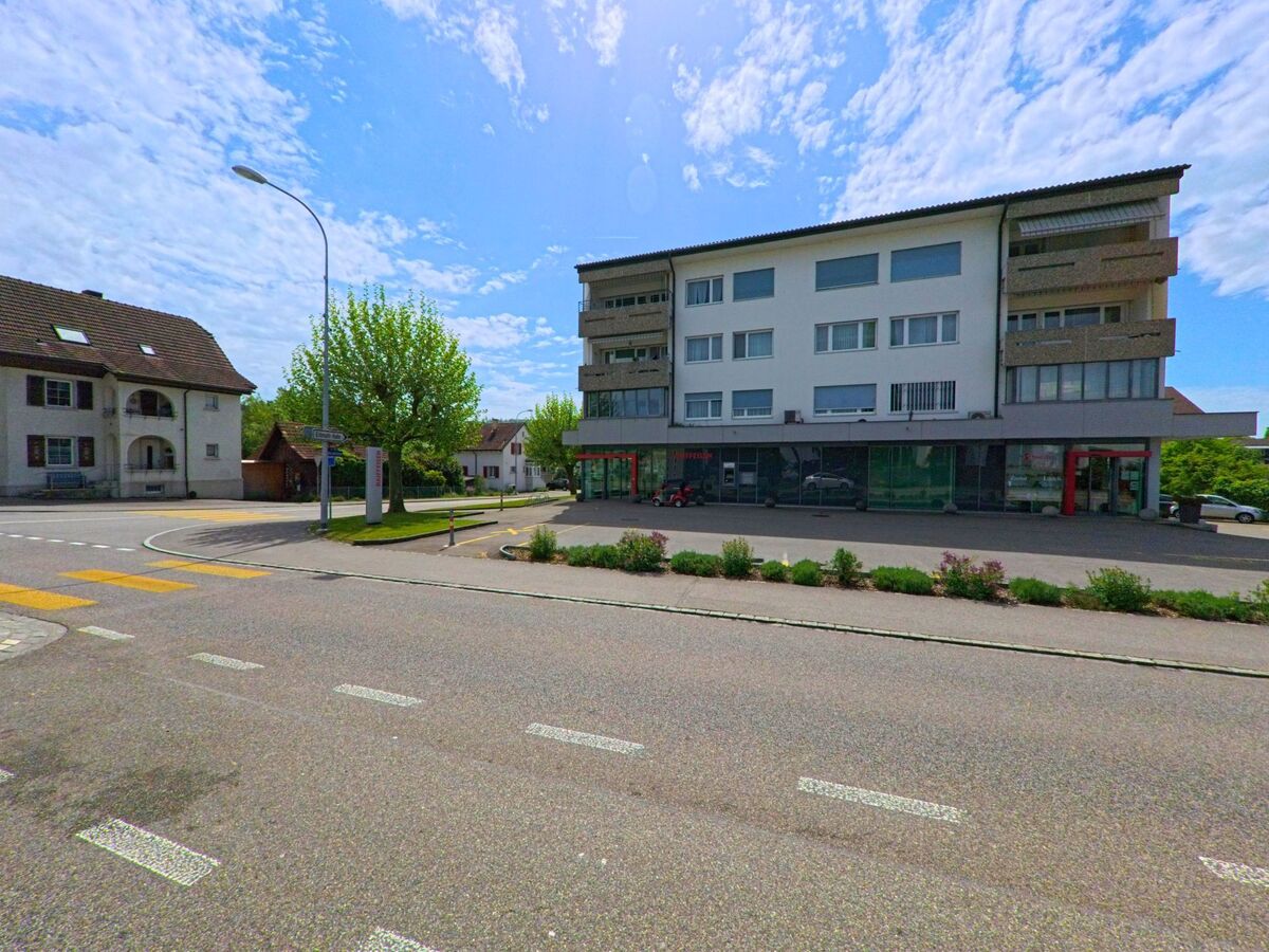  In Däniken (4658, Kanton Solothurn) steht ein modernes Wohn- und Geschäftshaus mit grossen Schaufenstern an einer Kreuzung mit Zebrastreifen.