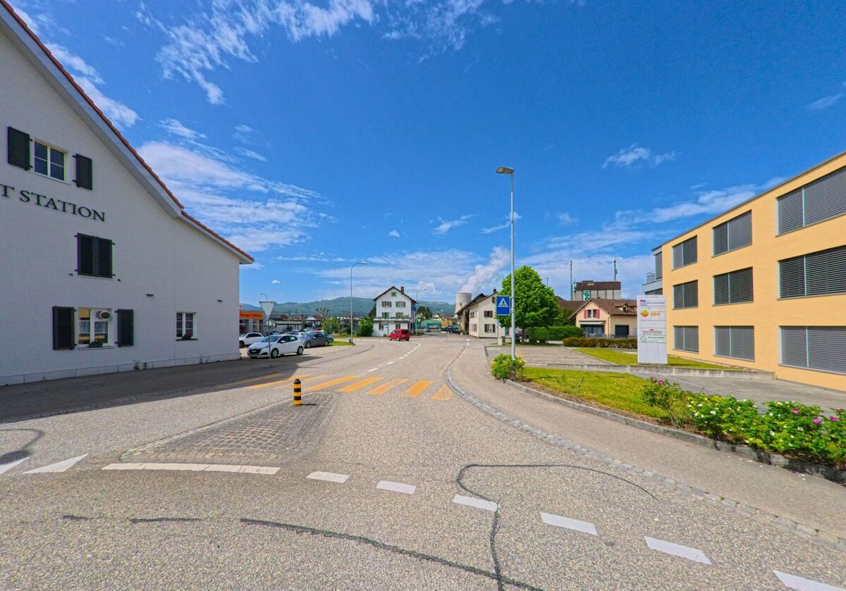 Eine zentrale Strassenszene in Däniken (4658, Kanton Solothurn) zeigt einen Zebrastreifen, moderne Bürogebäude und ein Restaurant mit Blick auf die nahen Hügel.